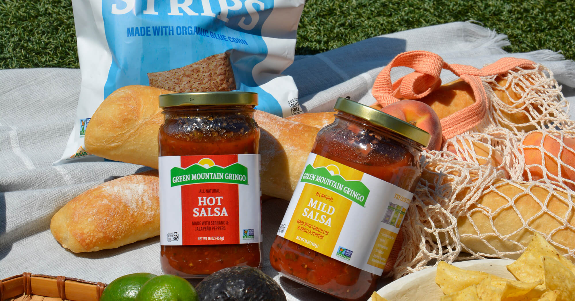 Jars of Green Mountain Gringo Salsa on a picnic blanket with other items such as bread and fruit.